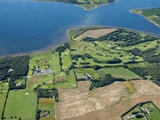 Luftfoto af en grøn golfbane med sandbunkere, omgivet af marker, træer, spredte bygninger og tæt på et stort blåt vandområde.
