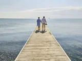 To mennesker går side om side på en træmole, der strækker sig ud over roligt havvand, på vej mod horisonten under en klar himmel.