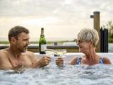 En mand og en kvinde slapper af i et boblebad, mens de klirrer med champagneglassene og smiler til hinanden, med en flaske champagne i nærheden og en naturskøn udendørs baggrund.