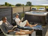 To personer i morgenkåber slapper af i liggestole ved et udendørs spabad og læser bøger i en solrig, indhegnet have med en naturskøn udsigt over marker og havet i baggrunden.
