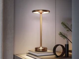 En moderne bordlampe i guld står på en stak bøger ved siden af en lille plante og et stearinlys på en lys overflade i et minimalistisk rum.