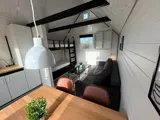 Et lille, moderne opholdsrum med en køjeseng, en grå sofa, et sofabord i glas og et spisebord med to stole, med hvide vægge og sorte loftsbjælker.