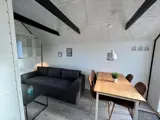 Et lille, moderne opholdsrum med en mørk sofa, et sofabord af glas, et spisebord af træ med fire stole, en potteplante og vægmonterede hylder under et skråt loft med synlige bjælker.