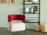 En moderne stol med rødt ryglæn og lys pude står ved siden af en sort hylde med bøger, indretning og kunstartikler; to flettede kurve står på gulvet i nærheden.