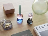 En bordplade med farverige glaslysestager, et tændt blåt lys, bøger, en glasbeholder med guldgenstande, en grøn vase og en lukket kasse på en lys træoverflade.
