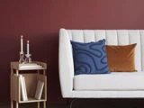 En moderne stue med en hvid sofa, blå og orange puder, et sidebord i træ, bøger og to tændte stearinlys mod en vinrød væg.