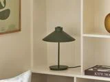 En moderne, mørkegrøn bordlampe står på en hvid hylde ved siden af en bog og pyntegenstande i en minimalistisk bolig.