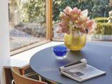 På et rundt bord ved et vindue står en gul vase med lyserøde blomster, en blå glasskål og en sammenrullet avis. Sollyset strømmer ind gennem haven udenfor.