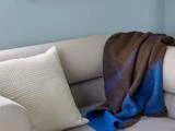 En cremefarvet pude og et brunt og blåt tæppe er placeret på en lysegrå sofa mod en lyseblå væg.