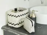En flettet kurv, en lille toilettaske og en sæbedispenser er placeret på en mørk bordplade ved siden af en hvid vask.