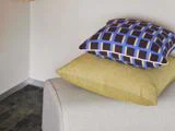To puder på en lysegrå sofa; den øverste pude har et geometrisk mønster i blåt, sort og gult, og den nederste pude er ensfarvet gul.