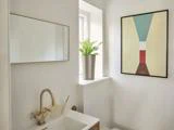 Et moderne badeværelse med et rektangulært spejl, potteplante i vindueskarmen, abstrakt vægkunst og en vask med guldarmatur.