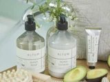 To flasker af mærket ALTUM med mærkerne Håndsæbe og Håndlotion, en tube håndcreme, en avocado og en træbørste på en bakke med blomster i baggrunden.