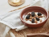 En brun skål med yoghurt toppet med brombær, mandler og granola står på en vævet måtte ved siden af en hvid serviet og en træske.