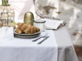 Et bord med to croissanter på en tallerken, et krus, en gaffel og en kniv, et dekorativt æg og en åben bog, alt sammen på en hvid dug.