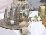 Bord med en glaskop med bladmønster, trææg, en lille vase med blomster, en guldbeholder, kurvebakke og hvid stofserviet på en hvid flade.