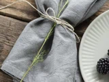 Grå stofserviet bundet med sejlgarn og en kvist, placeret på et rustikt træbord ved siden af en hvid tallerken med brombær på kanten.