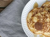 Tre pandekager med sirup på en hvid tallerken, placeret ved siden af en grå stofserviet på et træbord.