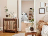 Hyggelig stue med neutrale toner, rustikke træmøbler, potteplanter, indrammet kunst og udsigt til et blødt oplyst soveværelse med en seng og en trækommode.