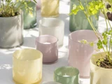 Forskellige pastelfarvede glaslysestager og urtepotter med grønne planter er arrangeret på en hvid flade i naturligt sollys.