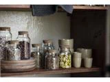 Glaskrukker med korklåg med forskellige bønner og krydderier står på en træhylde sammen med stablede keramiske kopper og skåle.
