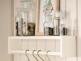 En hvid væghylde rummer glaskrukker med tørrede krydderurter eller krydderier, et indrammet blomsterprint og har flere metalkroge hængende under sig.