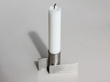 Et hvidt stearinlys står oprejst i en moderne, sølvfarvet metalholder med geometriske, rektangulære former og kaster en svag skygge på en lys overflade.