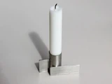 Et hvidt stearinlys står oprejst i en moderne, sølvfarvet metalholder med geometriske, rektangulære former og kaster en svag skygge på en lys overflade.