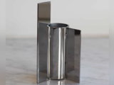 En moderne, metallisk vase med en cylindrisk midte og to flade, lodrette paneler på hver side, placeret på en marmoroverflade.