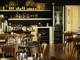 Et hyggeligt restaurantinteriør med træborde og -stole, dækket med glas og bestik. På hylderne står vinflasker og glas, og i baggrunden ses et køleskab og en disk.