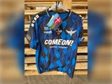 En blå og sort mønstret fodboldtrøje med sponsorlogoer hænger på en bøjle med et lille dansk flag fastgjort til den.