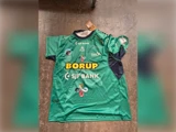 Grøn sportstrøje med korte ærmer, holdlogoer, sponsorer og en signatur nederst til venstre.
