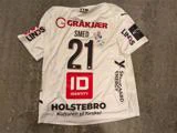 Hvid fodboldtrøje med nummer 21 og forskellige sponsorlogoer på ryggen, herunder en håndskrevet underskrift ved siden af nummeret.