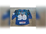 En blå og sort sportstrøje med påskriften »DILLING VICTOR 98« på ryggen, sponsorlogoer og et lille flag over navnet. Trøjen ligger fladt ud på en betonoverflade.