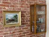 Et indrammet havmaleri hænger på en murstensvæg ved siden af et glasskab i træ med tallerkener, krus, glas og små krukker.