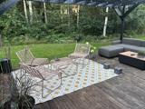 Udendørs terrasse med to kurvestole og et lille bord på et mønstret tæppe, en grå sektionssofa og grønne planter i baggrunden. Terrassen har trægulv og en pergola ovenover.