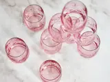 Ni lyserøde glasbægre med ætsede mønstre er arrangeret på en lys marmoroverflade, nogle stablet og andre placeret enkeltvis.