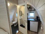 Et lille, moderne tekøkken med køleskab, vask, hylder og et spejl på døren fører til et kompakt badeværelse med toilet og håndvask. Vægge og loft er hvidmalet træ.