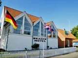 En hvid bygning med rødt tag med navnet Skagen Motel, med tyske, britiske, svenske og danske flag foran og et hvidt stakit under en klar blå himmel.