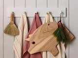 Et hvidt vægmonteret stativ med en lille kost, to køkkenhåndklæder, et træskærebræt i form af en gris og en dekorativ kvist af stedsegrønne blade.