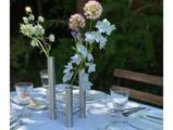 En moderne metalvase med tre rør rummer lyseblå og lilla blomster på et bord, der er dækket med et hvidt bordtæppe og klargjort til et måltid.