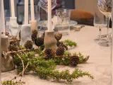 Nærbillede af et bord dækket med stearinlys i lysestager, fyrrekogler og grene af stedsegrønne træer samt glas og tallerkener, der tilsammen skaber en festlig og naturlig borddekoration.