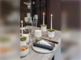 Et træbord dækket med tallerkener, bestik, en serviet, glas, småkager og fire hvide stearinlys i lysestager med mos, hvoraf nogle er tændt, hvilket skaber en hyggelig, rustik spiseatmosfære.