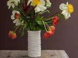 En hvid, ribbet vase rummer en buket af hvide og orange blomster med grønne blade, der står på et rustikt træbord foran en brun baggrund.