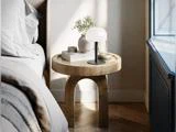 Et rundt natbord af træ med en lampe, bøger, en vase med blomster og en lille skål ved siden af en pænt redt seng i et lyst, moderne værelse.