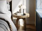 På et rundt natbord af træ står der en lampe, en lille vase med blomster, en bog og en skål ved siden af en seng med gråt sengetøj, tæt på et solbeskinnet vindue.