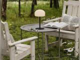 To træstole med hynder står ved et lille rundt bord, hvorpå der står en lampe, et blad, en bog og en tusindfryd, udendørs på en græsplæne med træer i baggrunden.