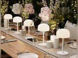 Et langt træbord dækket til et måltid, pyntet med hvide lamper, stearinlys, glas, tallerkener, druer samt lyserøde og hvide blomsterarrangementer.