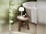 På en træskammel ved siden af badekarret står en hvid lampe og toiletartikler; i baggrunden ses grønne flisewægge og et håndklæde.