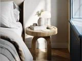 Et rundt natbord af træ med en hvid lampe, en vase med grønne planter, en bog og et dekorativt bakke ved siden af en redt seng og et vindue.